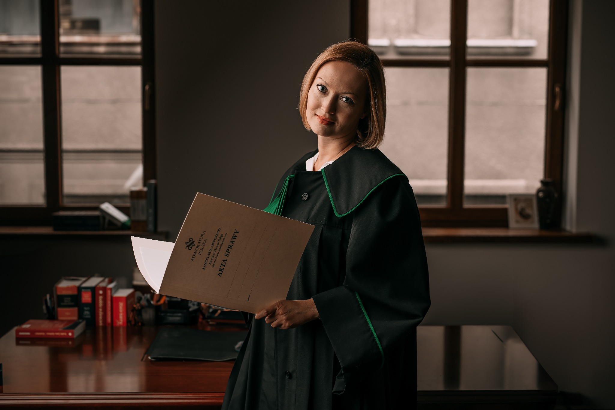Portret kobiety w todze adwokackiej, trzymającej teczkę z napisem 'Akta Sprawy', na tle biura z oknem.