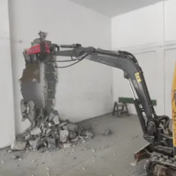 Mini koparka z hydraulicznym młotem pneumatycznym rozbija ścianę wewnątrz budynku, gruz leży na podłodze, widoczna drabina w tle.