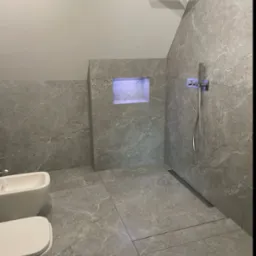 Nowoczesna łazienka w odcieniach szarości z marmurkową glazurą, kabina prysznicowa typu walk-in z odpływem liniowym, podwieszana toaleta i bidet, oraz podświetlana wnęka w ścianie.