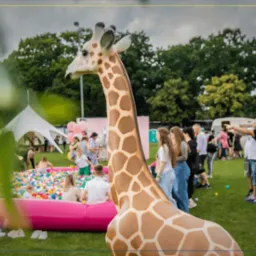 Plenerowa strefa zabaw z dmuchanym basenem wypełnionym kolorowymi piłeczkami, żyrafa dekoracyjna na pierwszym planie, w tle biały namiot i bawiący się ludzie.