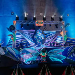 Scena z futurystyczną dekoracją, podświetlona niebieskim światłem, z geometrycznymi wzorami, sztucznymi kwiatami i postacią z neonowymi skrzydłami na pierwszym planie.