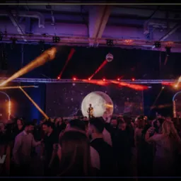 Impreza firmowa z saksofonistą na scenie przed ekranem z motywem księżyca, oświetlenie sceniczne, parkiet z bawiącymi się ludźmi.