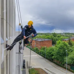 Pracownik w kasku i uprzęży, zawieszony na linach, czyści elewację budynku za pomocą myjki ciśnieniowej; w tle widok na miasto z kominem i pochmurne niebo.