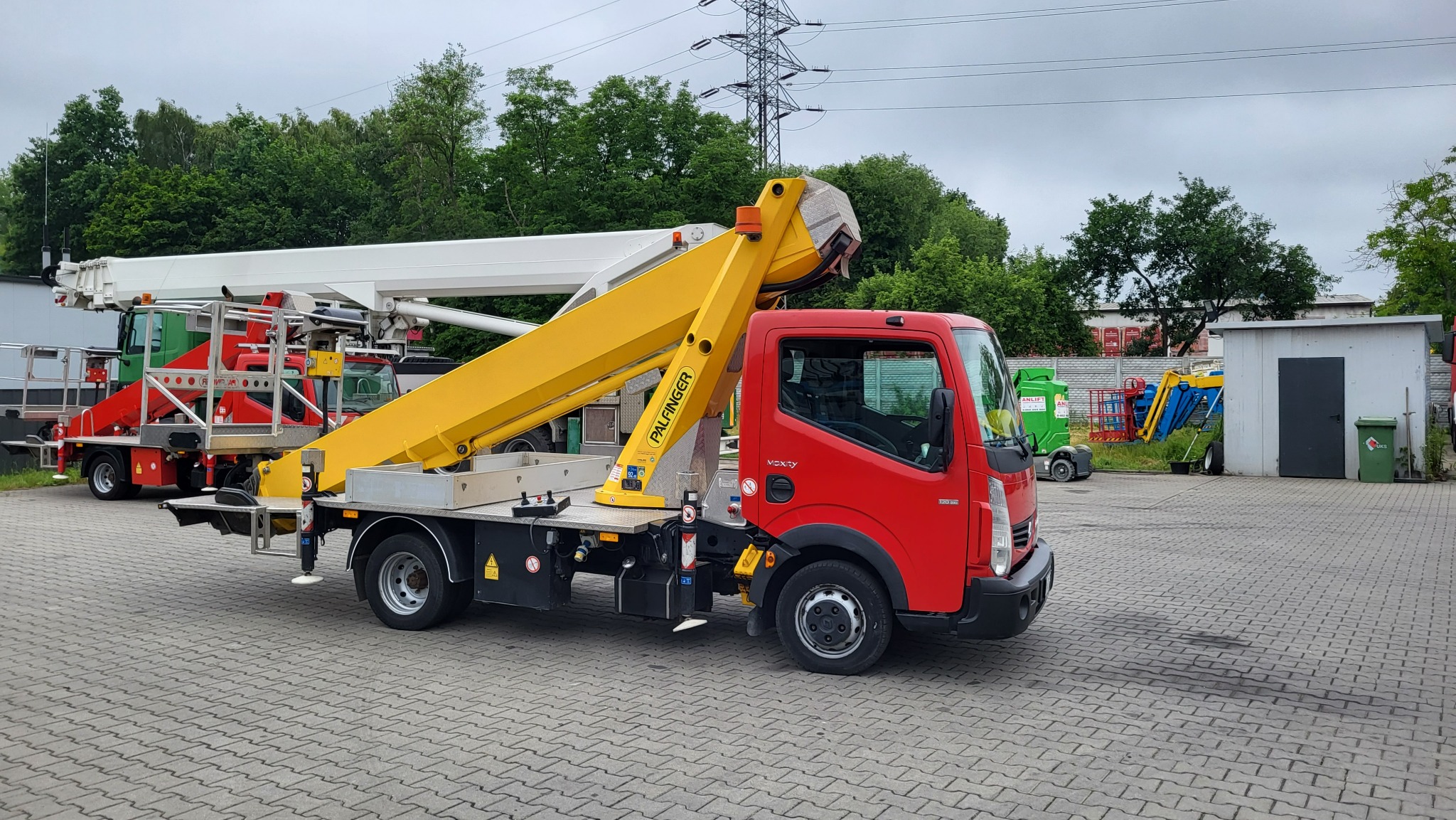 Czerwony samochód z żółtym podnośnikiem koszowym Palfinger na parkingu z kostki brukowej, w tle zieleń drzew i słupy wysokiego napięcia.