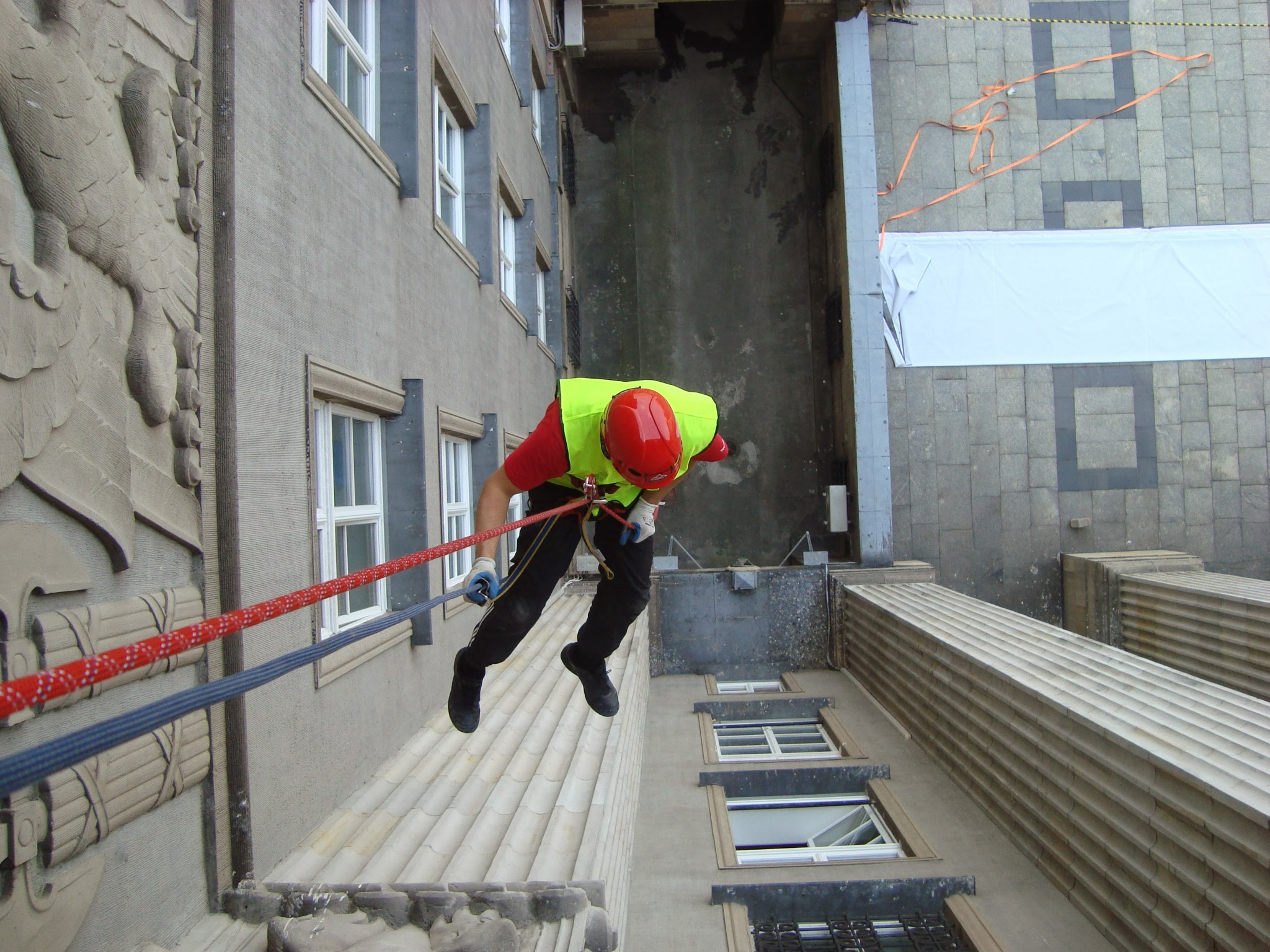 Pracownik w kasku i kamizelce odblaskowej, zwisający na linach z wysokości budynku z ozdobną elewacją, widok z góry.