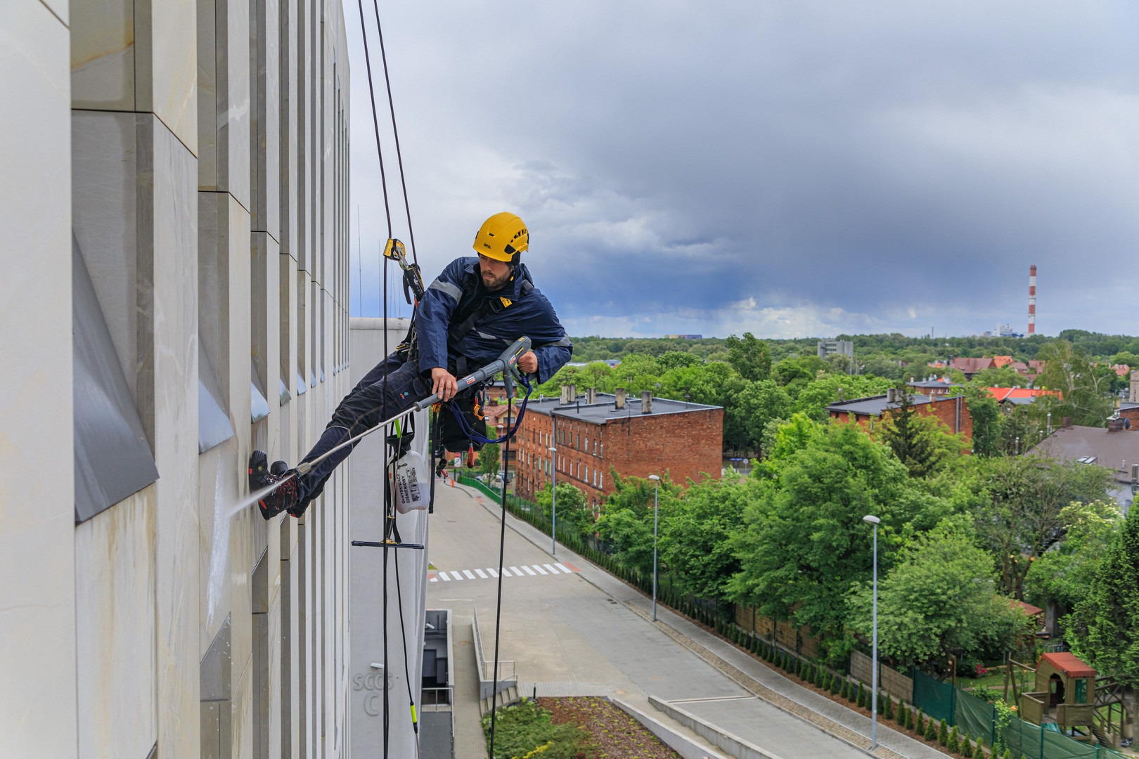 Pracownik w kasku i uprzęży, zawieszony na linach, czyści elewację budynku za pomocą myjki ciśnieniowej; w tle widok na miasto z kominem i pochmurne niebo.