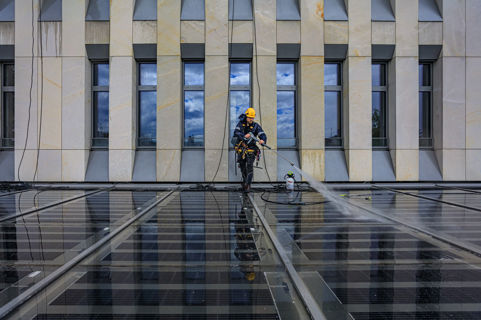 Pracownik w kasku i uprzęży, z myjką ciśnieniową, czyści szklany dach budynku o nowoczesnej architekturze, z widocznymi refleksami nieba w oknach.
