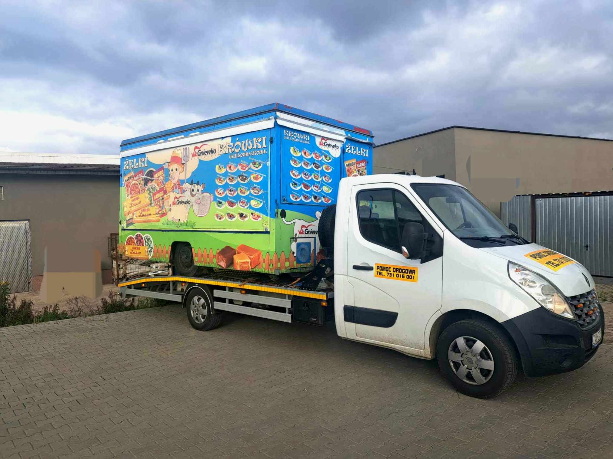 Biała autolaweta z żółtymi pasami transportuje furgonetkę z kolorową reklamą słodyczy, w tle budynek i pochmurne niebo.