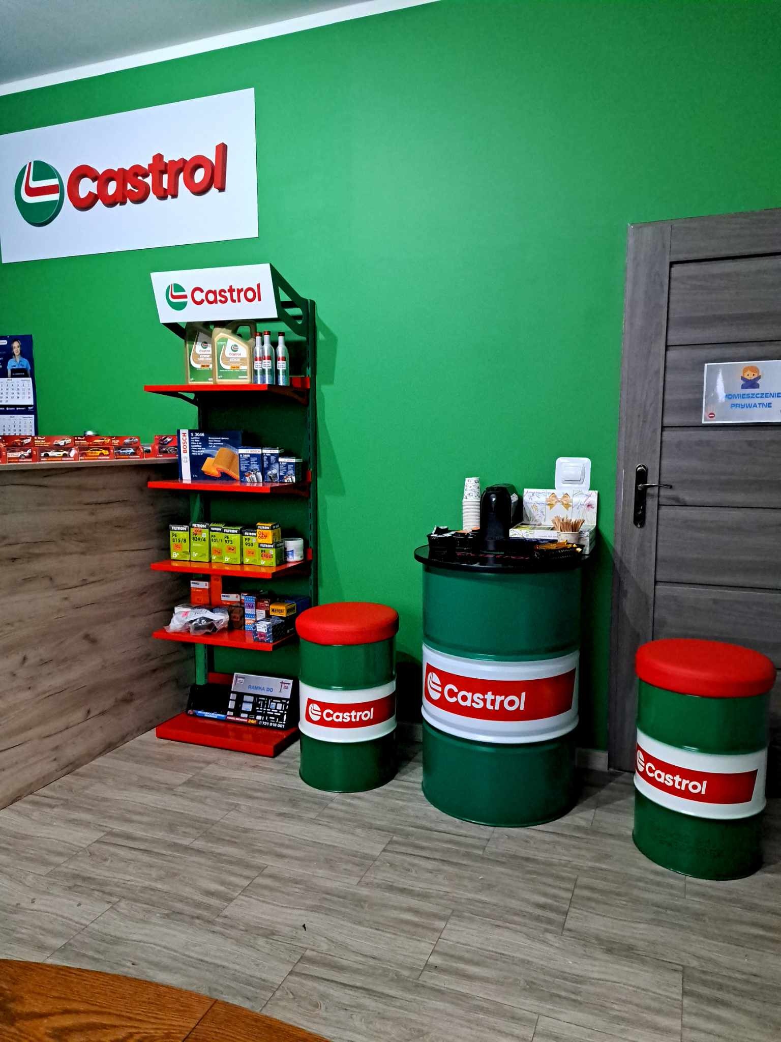 Wnętrze warsztatu samochodowego z zielonymi ścianami, regałem z olejami i filtrami marki Castrol, oraz stolikami/pufami wykonanymi z beczek z logo Castrol, przy drzwiach tabliczka 'Pomieszczenie...