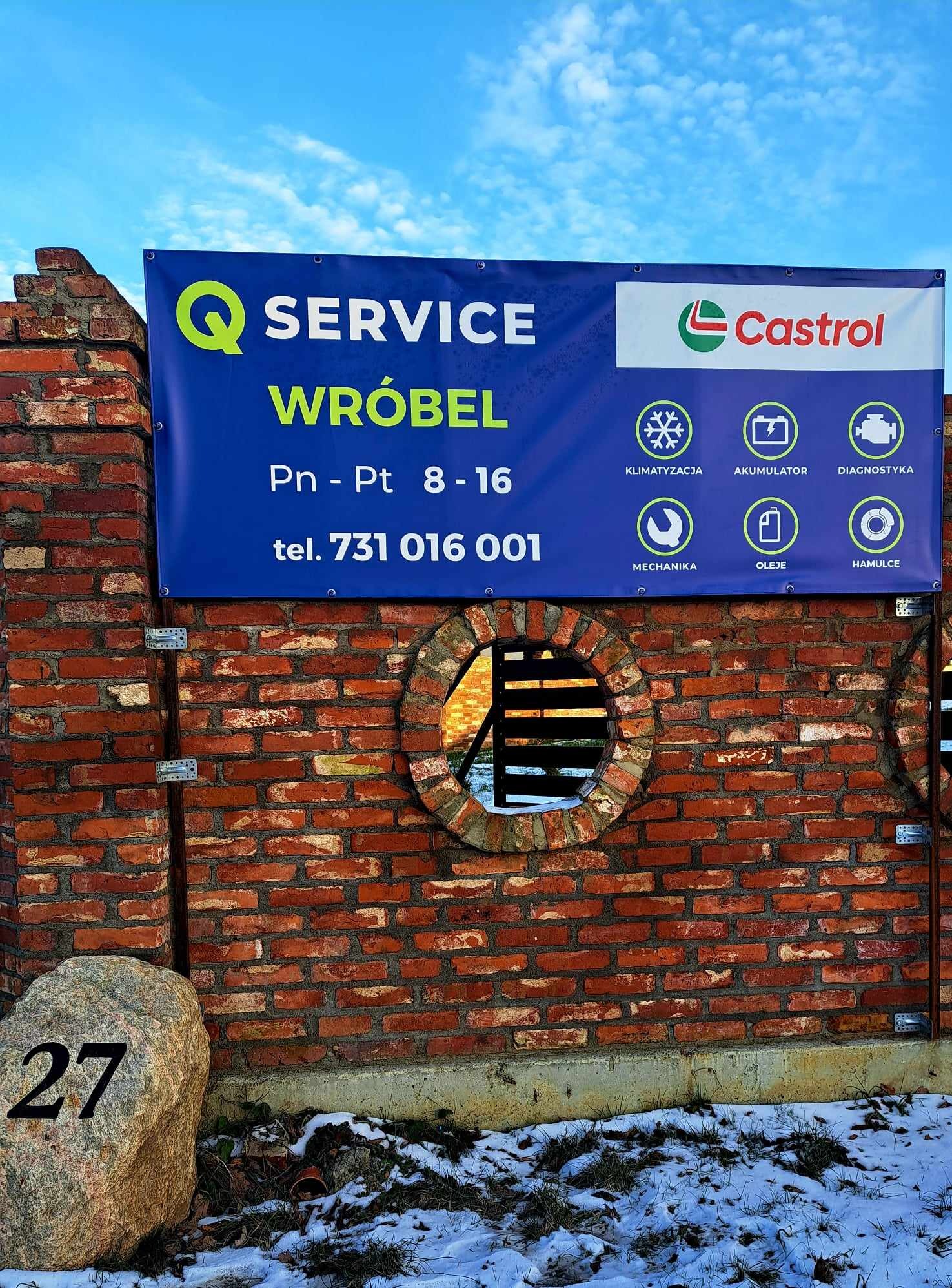 Tablica reklamowa serwisu samochodowego 'WRÓBEL' z logo Castrol, umieszczona na ceglanym murze z okrągłym otworem, obok kamienia z numerem 27.
