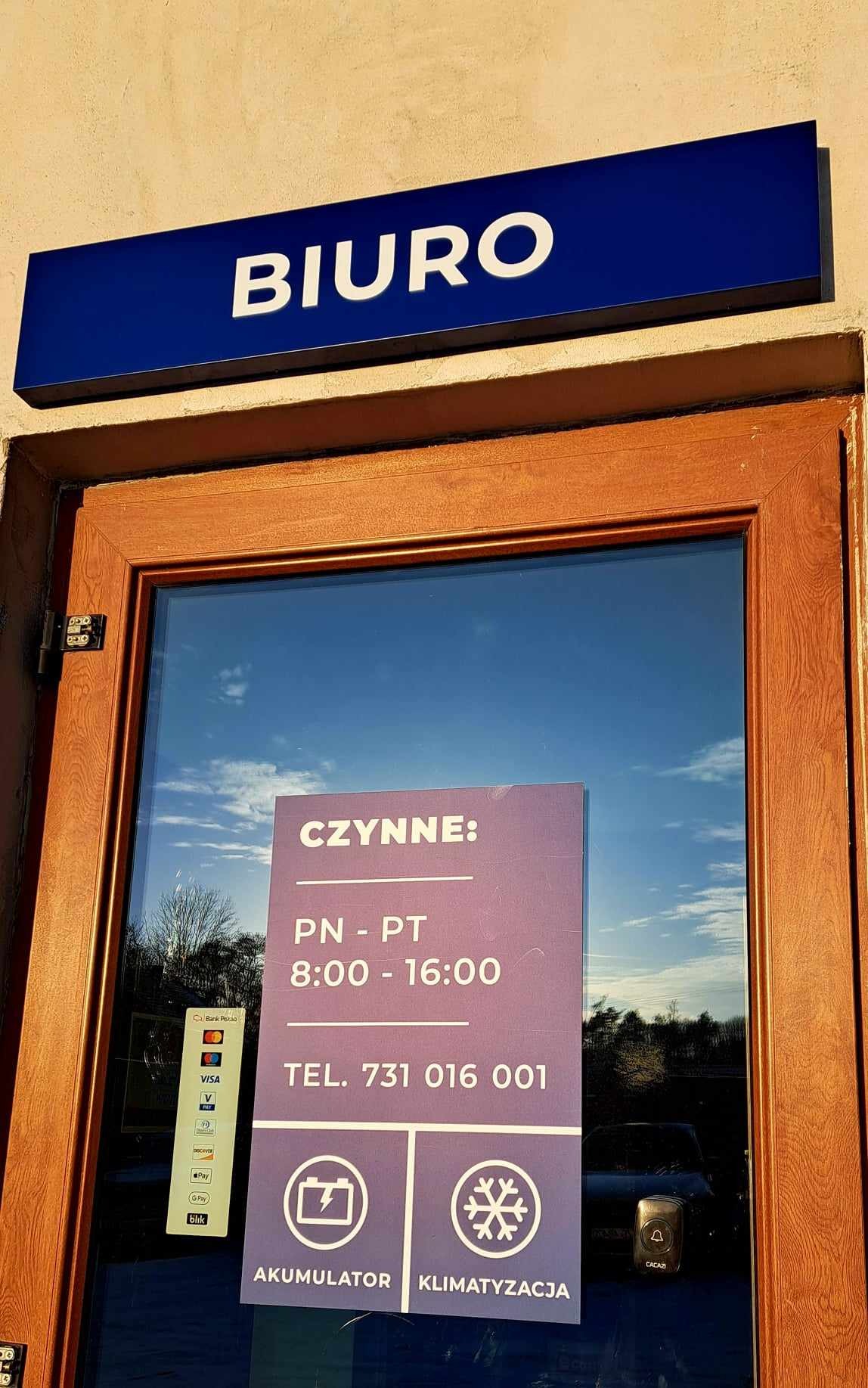 Wejście do biura z szyldem 'BIURO', informacją o godzinach otwarcia, numerem telefonu oraz akceptowanych metodach płatności, w tym płatności kartą i BLIK. Na drzwiach naklejka z ikonami akumulatora...
