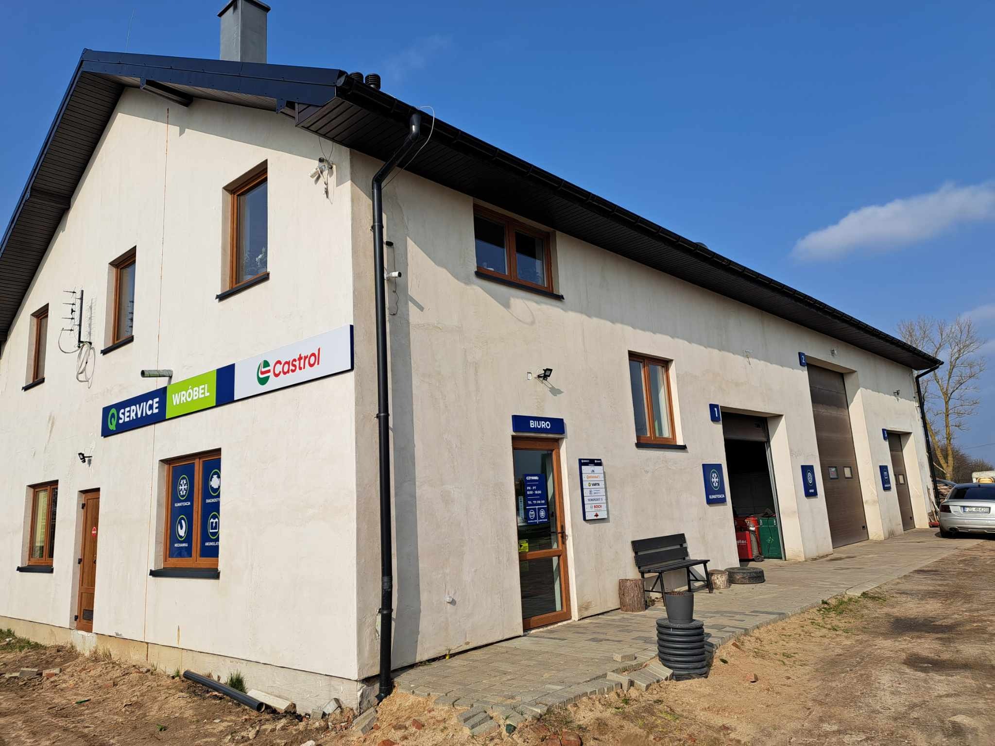 Zewnętrzny widok budynku warsztatu samochodowego z logo firm Castrol i Service Wróbel, jasna elewacja, czarny dach i okna z brązowymi ramami.