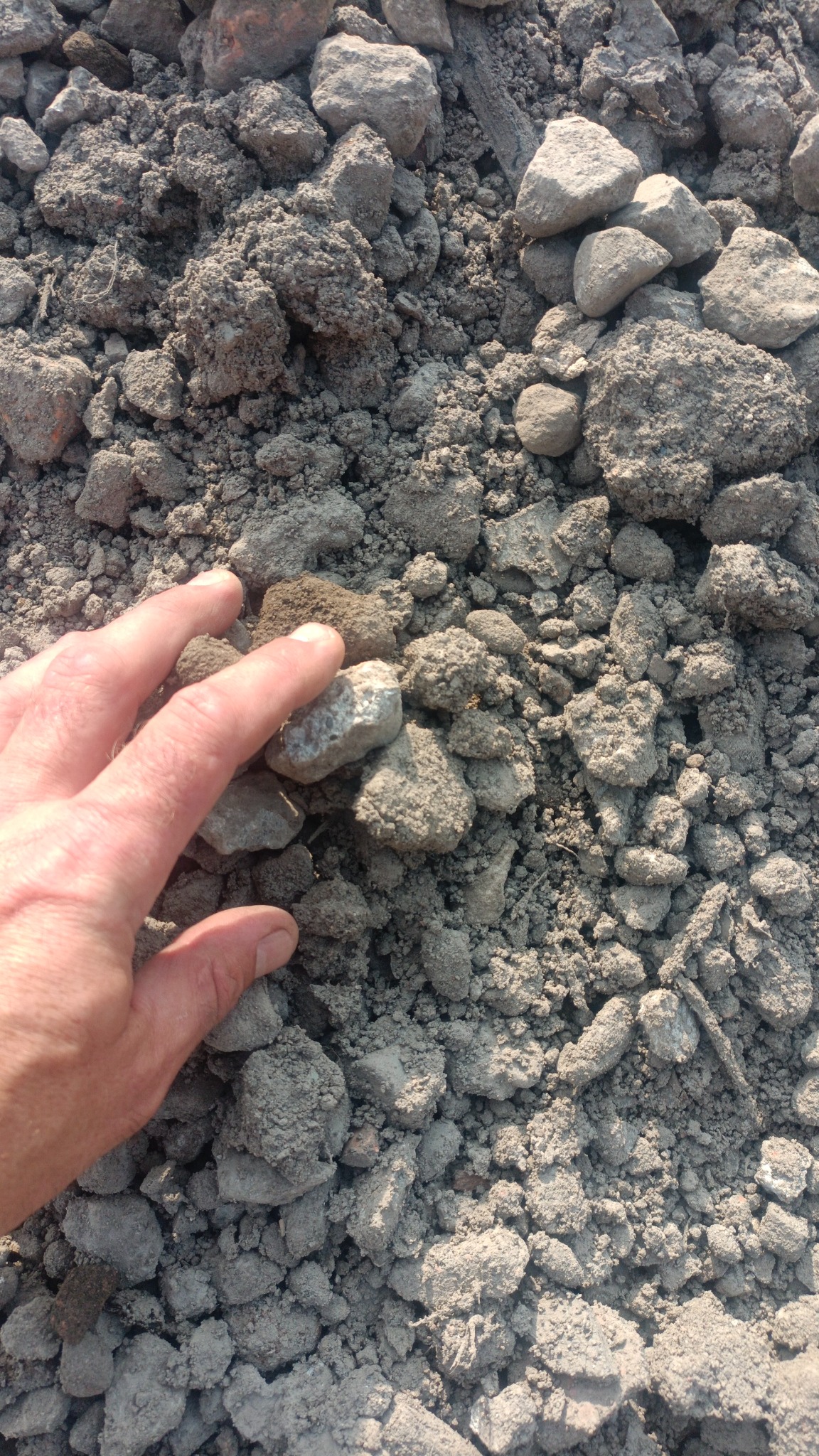 Dłoń dotyka grudek ziemi i kamieni o szarej barwie, prawdopodobnie podczas prac ziemnych.