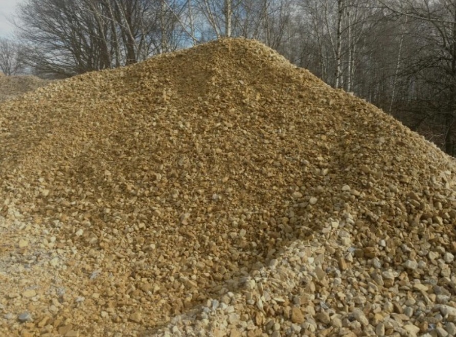 Kupa żwiru o zróżnicowanej granulacji w odcieniach beżu i brązu, z widocznymi drzewami bezlistnymi w tle.