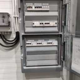 Otwarta szara skrzynka rozdzielcza z widocznymi bezpiecznikami, wyłącznikami i oznaczeniami 'Wył. Główny', 'Zab. Ochronnika', 'Ochronnik' oraz 'Zest. Gniazd', zamontowana na ścianie z panelami. Po...