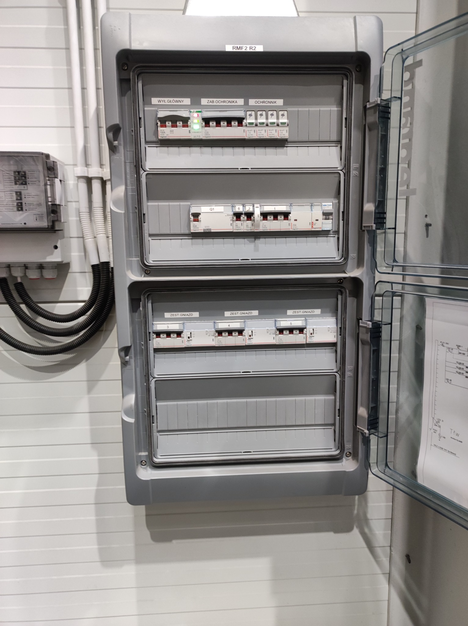 Otwarta szara skrzynka rozdzielcza z widocznymi bezpiecznikami, wyłącznikami i oznaczeniami 'Wył. Główny', 'Zab. Ochronnika', 'Ochronnik' oraz 'Zest. Gniazd', zamontowana na ścianie z panelami. Po...