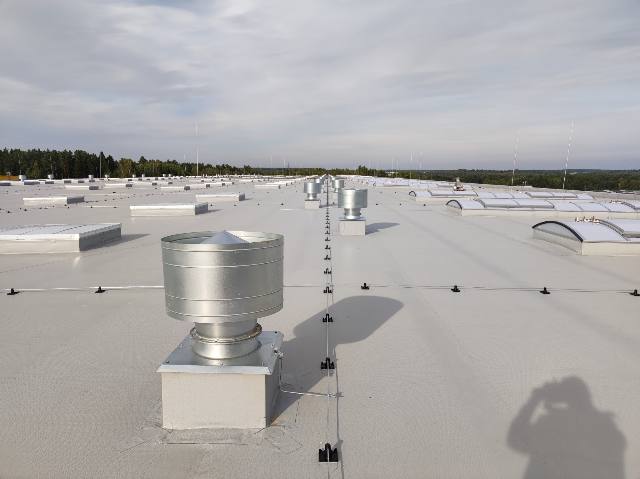 Widok z góry na płaski dach przemysłowy z licznymi świetlikami, kominami wentylacyjnymi i systemem odgromowym z przewodami i uchwytami; w tle widoczny las.