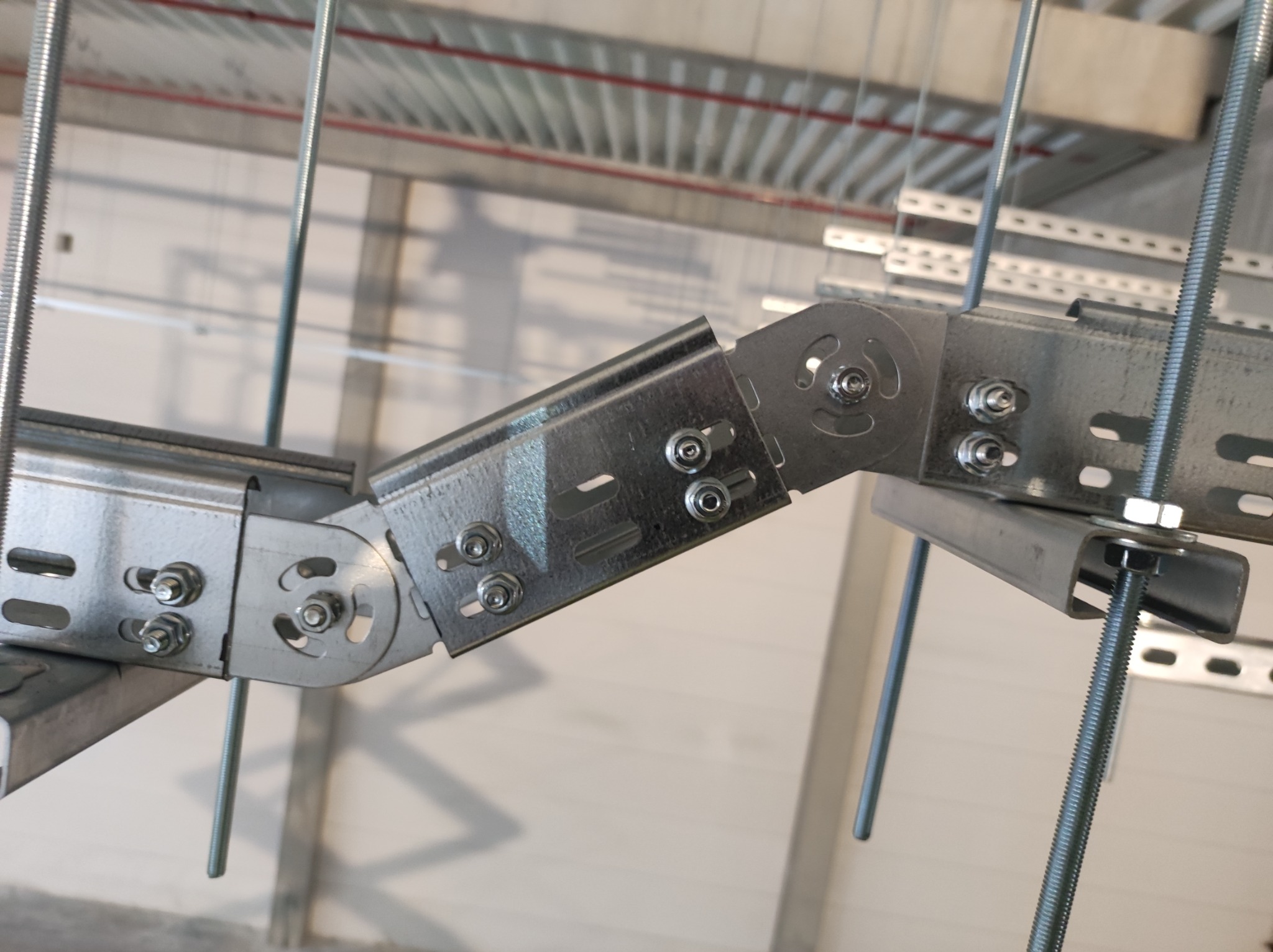 Metalowa konstrukcja wsporcza z perforowanych profili i śrub, zawieszona pod sufitem hali przemysłowej, widoczne pręty gwintowane i elementy złączne.