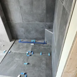 Układanie ciemnoszarych płytek wielkoformatowych na podłodze i ścianach z użyciem niebieskich klinów poziomujących, widoczny profil wykończeniowy przy drzwiach.