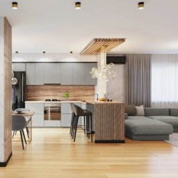 Komarova Design - Nowoczesna kuchnia z szarymi szafkami, drewnianym panelem nad blatem i wyspą z drewnianymi listwami, oświetlona wiszącą lampą w kształcie kuli, z widocznym fragmentem salonu z szarą sofą narożną.