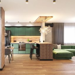 Komarova Design - Nowoczesna kuchnia w odcieniach zieleni z drewnianym blatem i wyspą, oświetlona dekoracyjną lampą wiszącą z kulistych kloszy, połączona z salonem z zieloną sofą modułową.