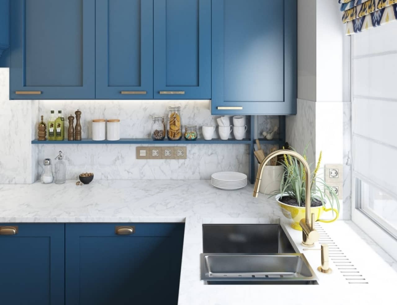 Niebieska zabudowa kuchenna z marmurowym blatem, złota armatura, rolety rzymskie w oknie, dodatki dekoracyjne na półce.
