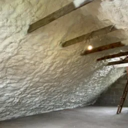 Poddasze ocieplone pianką PUR, widoczne drewniane belki stropowe i faktura pianki. Na suficie żarówka. Izolacja wykonana natryskowo, pokrywająca całą powierzchnię.