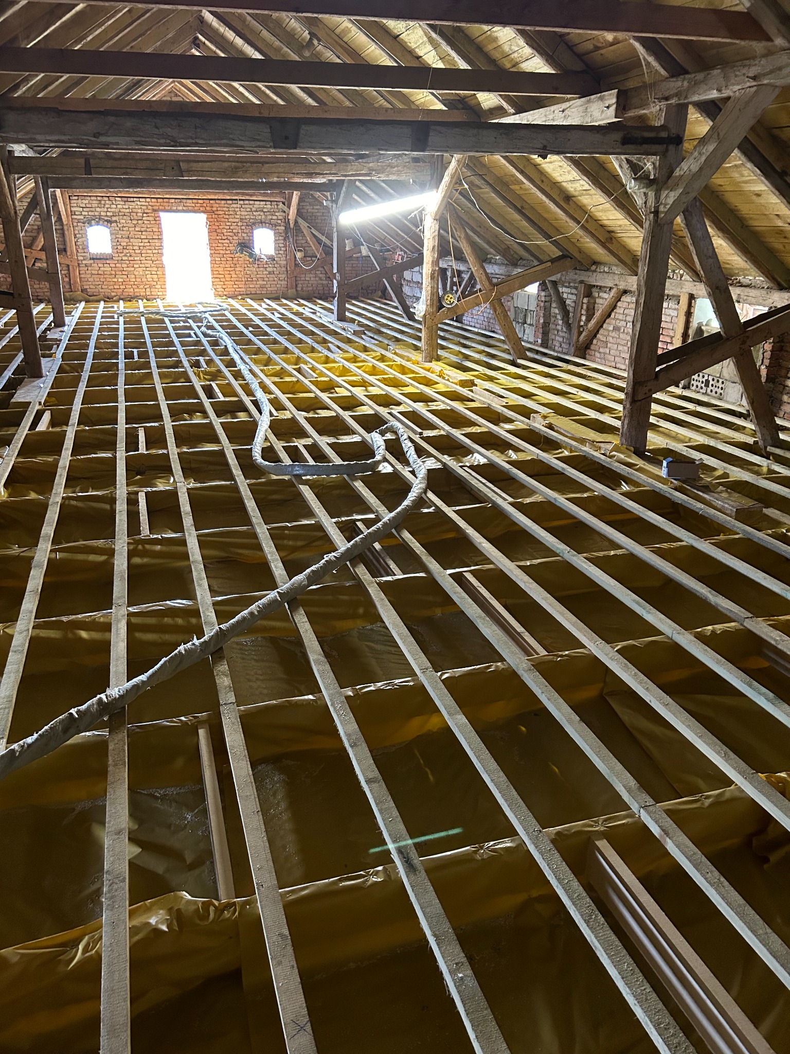 Konstrukcja drewniana poddasza zabezpieczona folią w kolorze złotym, widok z dołu na krokwie i deski, w tle ceglana ściana z oknami oraz wpadające światło słoneczne.