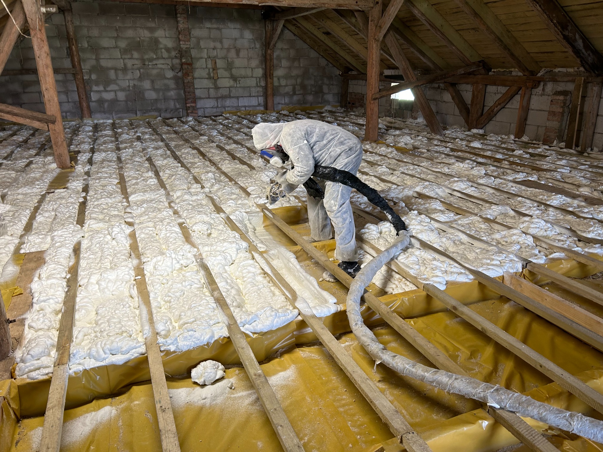 Pracownik w kombinezonie ochronnym aplikuje piankę PUR na poddaszu, widoczne drewniane belki konstrukcyjne i żółta folia zabezpieczająca, częściowo pokryte białą pianą.
