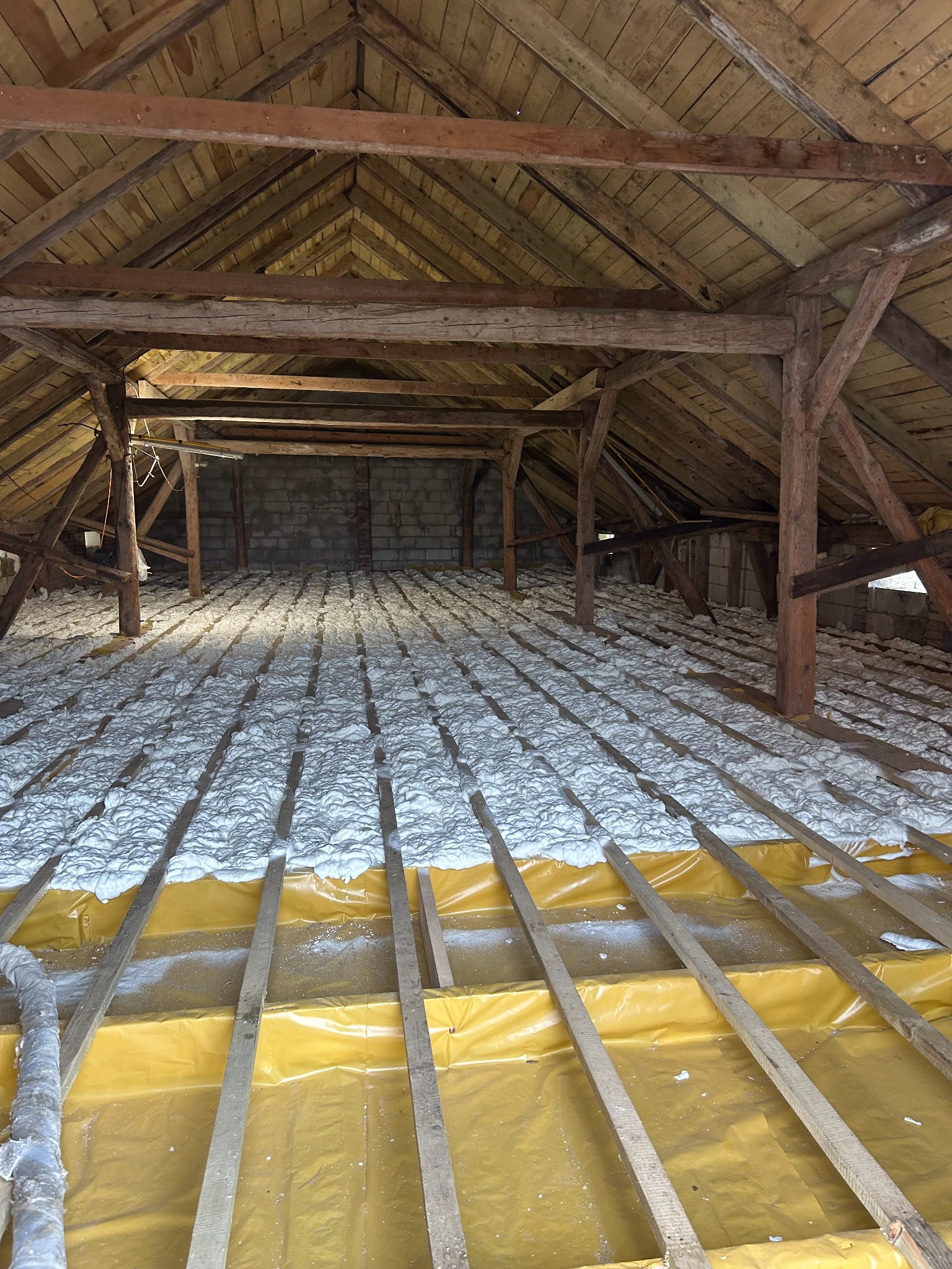 Poddasze z drewnianą konstrukcją pokryte żółtą folią i izolacją piankową, widoczne drewniane belki stropowe i fragment ściany z pustaków.
