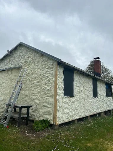 Dom po ociepleniu pianką PUR, widoczna drabina i stolik. Ściany pokryte grubą warstwą izolacji, okna zabezpieczone folią. Trawa i pochmurne niebo w tle.