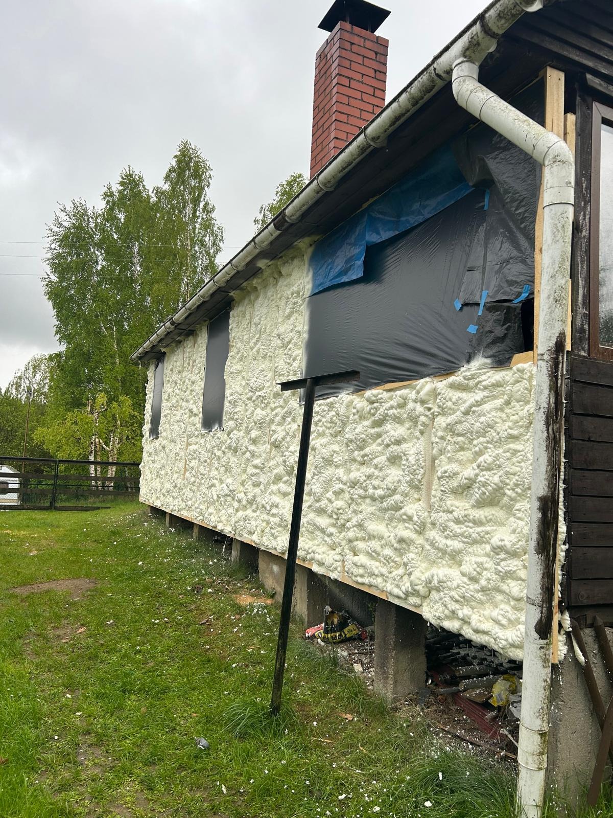 Ocieplanie ścian domu pianką PUR, widoczne zabezpieczone okna folią i fragment ceglanego komina. Prace w toku, budynek otoczony zielenią.