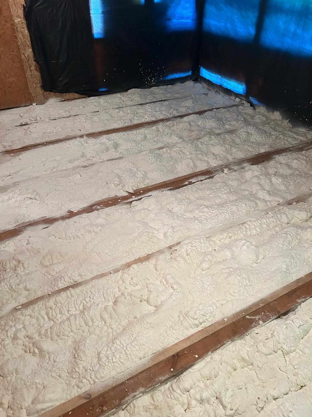 Poddasze po ociepleniu pianką PUR, widoczne drewniane belki i warstwa izolacji. Czarna folia w rogu pomieszczenia.