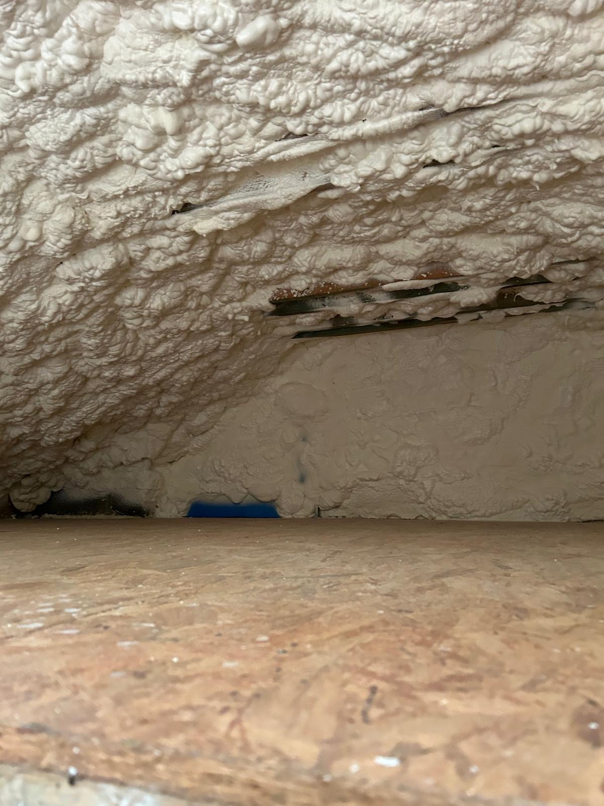 Poddasze po ociepleniu pianką PUR, widoczne białe, nierównomierne wypełnienie przestrzeni między krokwiami i fragment podłogi z płyty OSB w dolnej części kadru.