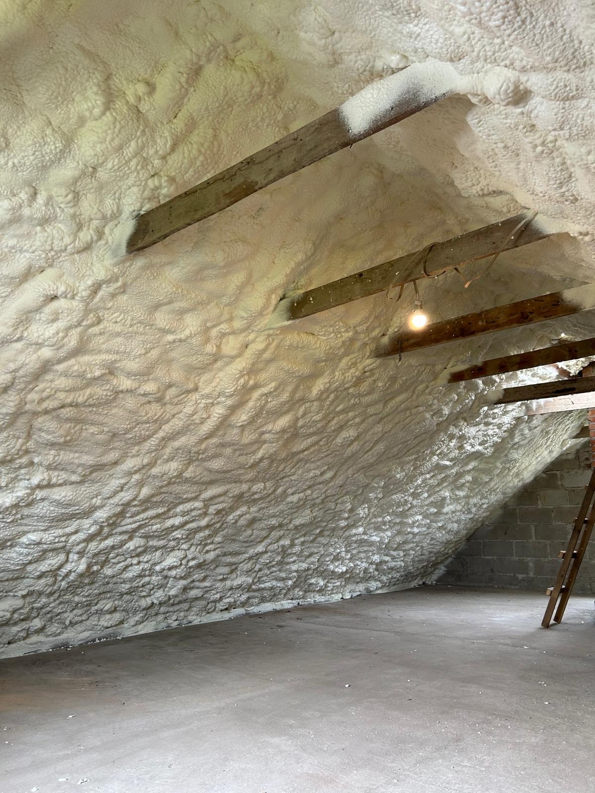 Poddasze ocieplone pianką PUR, widoczne drewniane belki stropowe i faktura pianki. Na suficie żarówka. Izolacja wykonana natryskowo, pokrywająca całą powierzchnię.