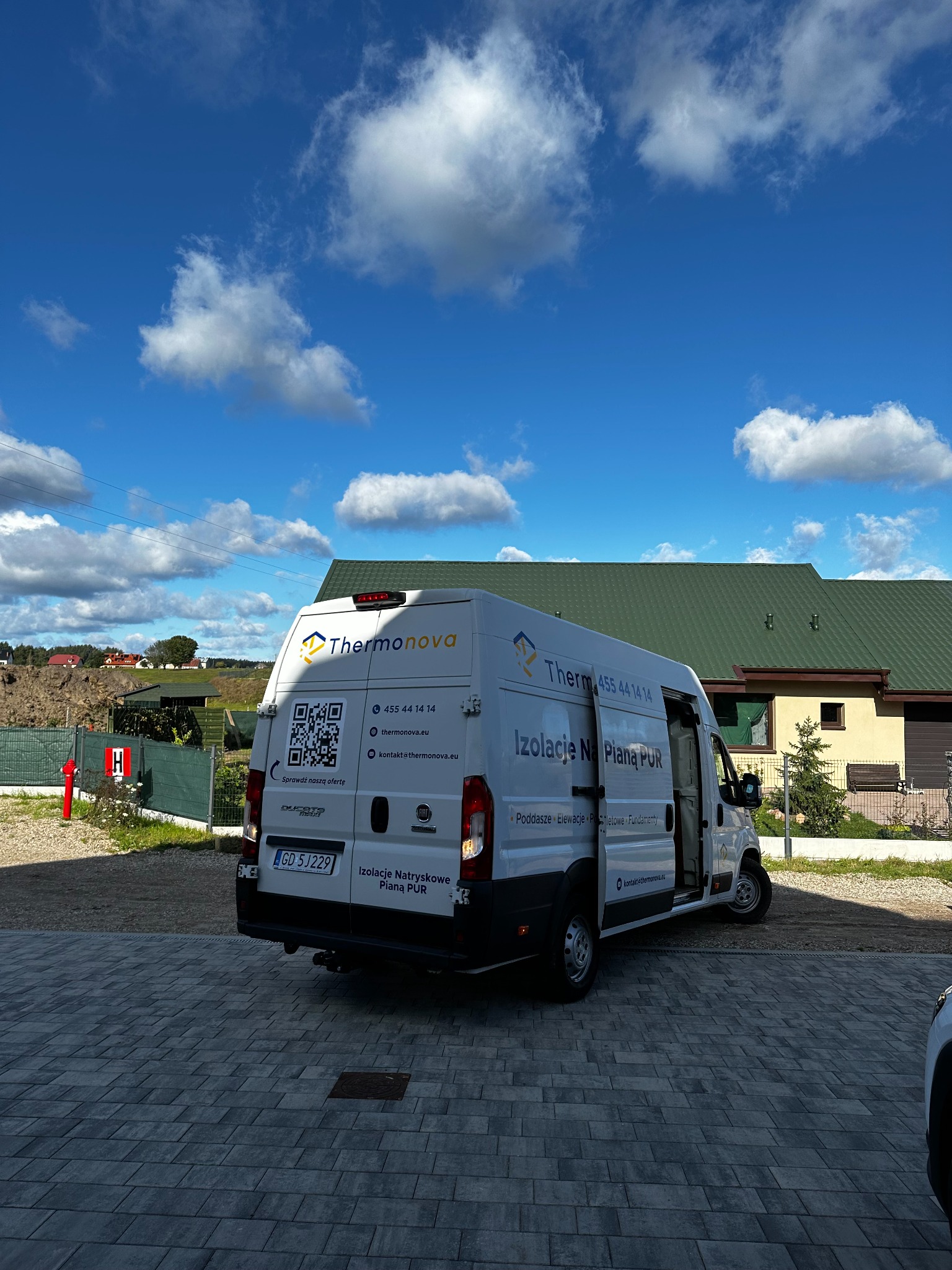 Biały samochód dostawczy firmy Thermonova z napisem 'Izolacje Natryskowe Pianą PUR' zaparkowany na brukowanym podjeździe przed domem z zielonym dachem, widoczne logo firmy, numer telefonu i adres...