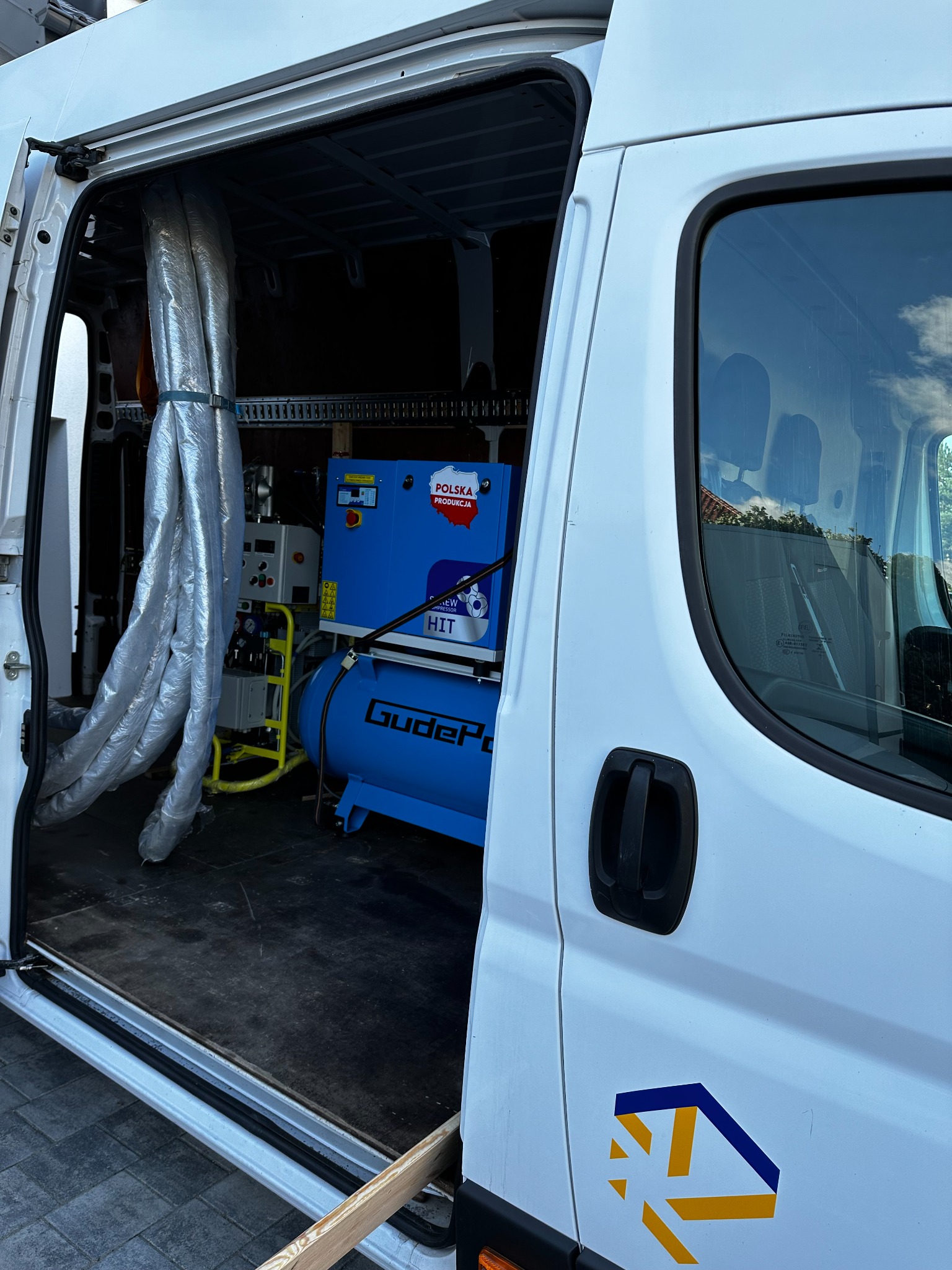 Wnętrze białego samochodu dostawczego z otwartymi drzwiami bocznymi, widoczny niebieski kompresor z napisem 'GudeP' oraz zwinięte, zabezpieczone folią rury, prawdopodobnie sprzęt do natrysku pianki.