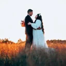 Para młoda w objęciach na tle zachodzącego słońca, wysoka trawa w kolorach złota i pomarańczy, ona w sukni z długim rękawem i kwiatem we włosach, on w czarnym garniturze.