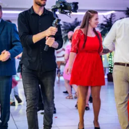 Kamerzysta z brodą i kolczykiem w uchu, filmuje parę w czerwonej sukience podczas wydarzenia, używając kamery z mikrofonem na stabilizatorze Ronin.