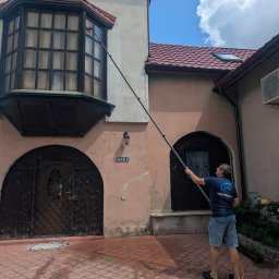 LTD Agnieszka Opioła C&M Clean and more - Mężczyzna czyści okno na wysokości za pomocą długiej lancy. Widoczny budynek z oknem typu wykusz i mokry bruk po czyszczeniu. Słoneczny dzień, błękitne niebo.