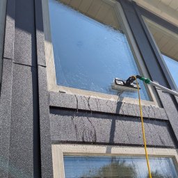 LTD Agnieszka Opioła C&M Clean and more - Mycie okna z zewnątrz za pomocą szczotki na teleskopowym kiju. Woda spływa po szybie. Widoczne elementy elewacji budynku i niebo.