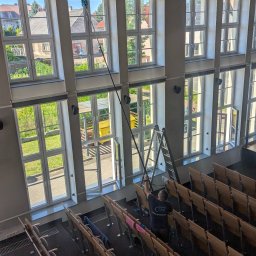 LTD Agnieszka Opioła C&M Clean and more - Pracownik myje wysokie okna w auli za pomocą teleskopowego kija z mopem. Aluminiowa drabina stoi obok. Widok z góry na rzędy krzeseł.