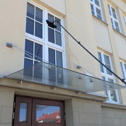 LTD Agnieszka Opioła C&M Clean and more - Mycie okien na wysokości w budynku z beżową elewacją. Widoczna lanca z szczotką i ślady wody po myciu okna. Ujęcie z dołu, na pierwszym planie przeszklone zadaszenie nad wejściem.