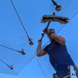 LTD Agnieszka Opioła C&M Clean and more - Mężczyzna na drabinie myje szklany daszek z metalowymi elementami mocującymi, w tle błękitne niebo i budynek. Widok z dołu, krople wody w powietrzu.