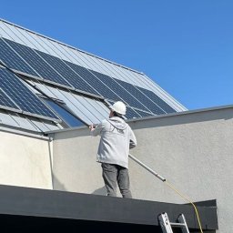 LTD Agnieszka Opioła C&M Clean and more - Pracownik w kasku czyści panele fotowoltaiczne na dachu budynku za pomocą długiej szczotki. Widoczna drabina i błękitne niebo.