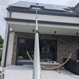 LTD Agnieszka Opioła C&M Clean and more - Czyszczenie paneli słonecznych na dachu domu jednorodzinnego przy użyciu teleskopowej szczotki. Widok z dołu na fasadę i panele w pochmurny dzień.