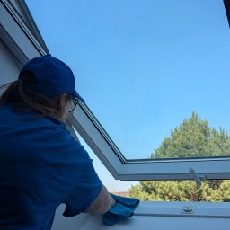 LTD Agnieszka Opioła C&M Clean and more - Osoba w niebieskiej koszulce czyści okno dachowe szmatką w Trzebini. Widok na błękitne niebo i drzewo. Skupienie na detalu czyszczenia ramy okiennej.