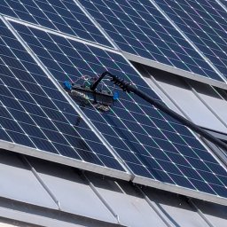 LTD Agnieszka Opioła C&M Clean and more - Czyszczenie paneli fotowoltaicznych na dachu budynku za pomocą specjalistycznej szczotki teleskopowej. Widoczne panele, dach z blachy i fragment ściany budynku.