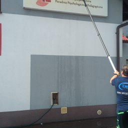 LTD Agnieszka Opioła C&M Clean and more - Pracownik myje elewację budynku z tablicą informacyjną 'Genius Loci' za pomocą długiego kija z myjką. Widoczny wąż doprowadzający wodę i mokra elewacja.