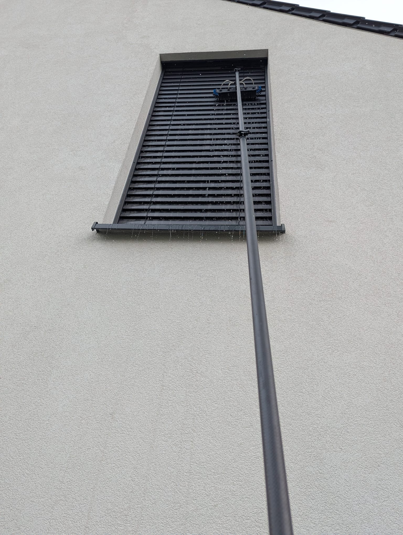 Mycie żaluzji zewnętrznych na elewacji budynku przy użyciu teleskopowej lancy z szczotkami. Woda spływa po lamelach.